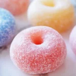 A plate of golden brown mochi donuts arranged in a circle, each donut topped with a glossy glaze and colorful sprinkles, resting on parchment paper.